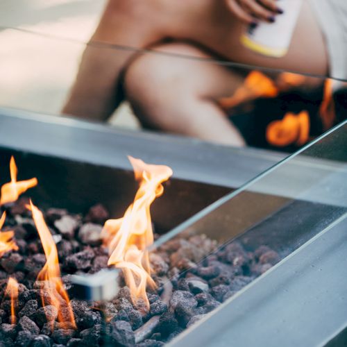 The image shows a person holding a canned beverage, sitting near a modern outdoor fire pit with glass sides and flames.