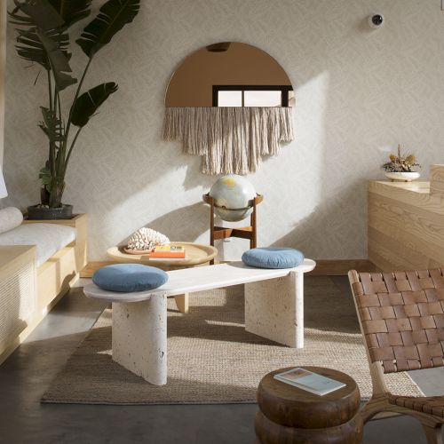 A cozy living room with a bench, marble table, two blue cushions, globe, wooden chair, rattan armchair, and decorative elements on a rug, ending the sentence.