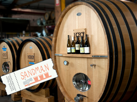 The image shows large wooden barrels in a winery, with wine bottles on a shelf attached to one of the barrels and a ticket labeled 