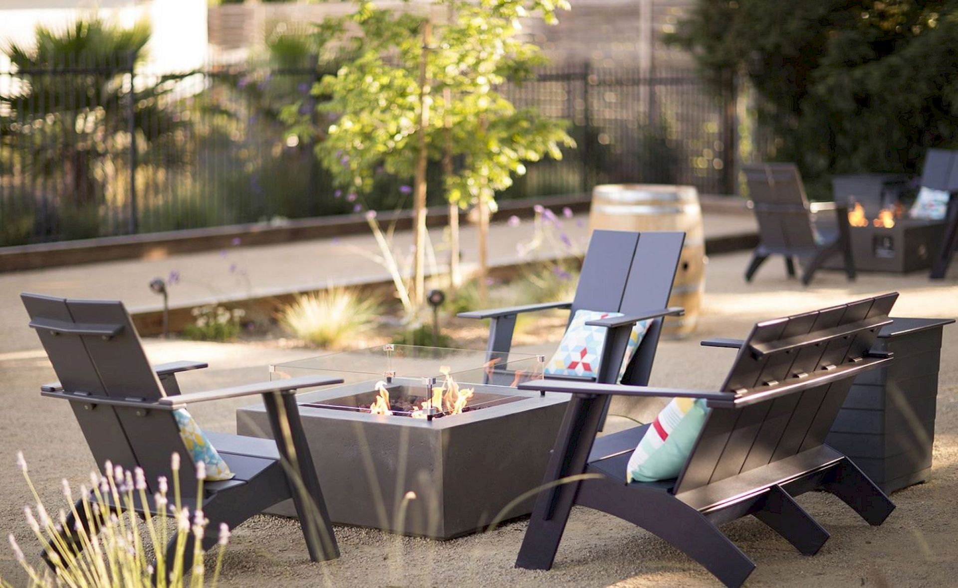 Outdoor seating area with Adirondack chairs around a fire pit, decorated with cushions, on a gravel surface.