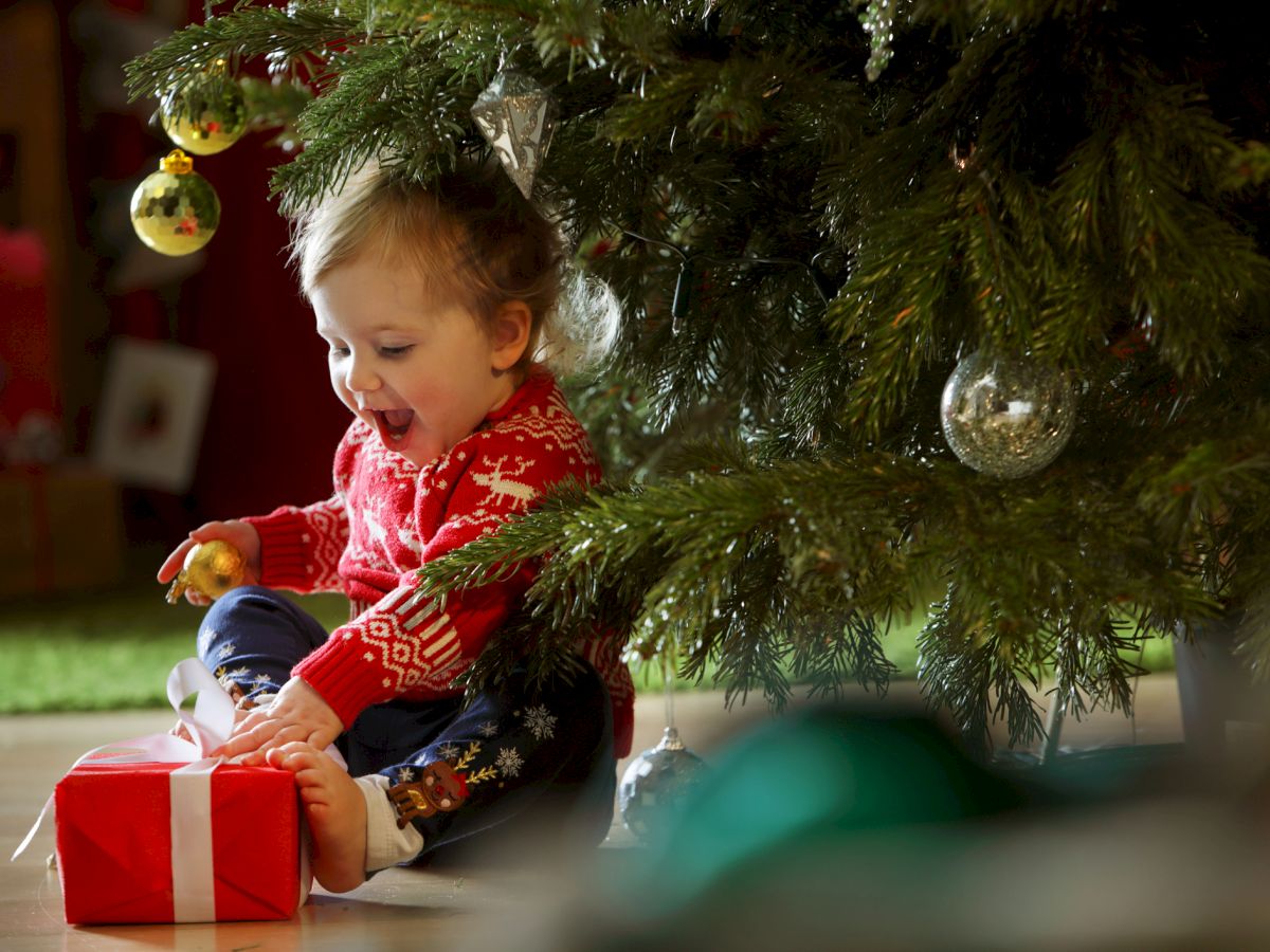 A child in a festive sweater is excitedly holding a wrapped gift under a decorated Christmas tree.