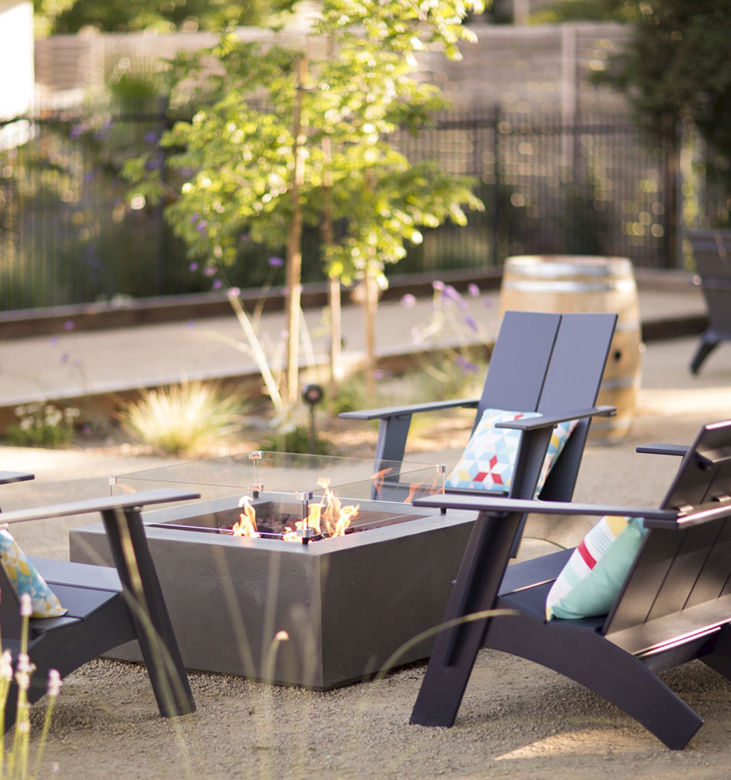 The image shows outdoor chairs surrounding a fire pit in a garden setting, with greenery and a decorative barrel in the background.