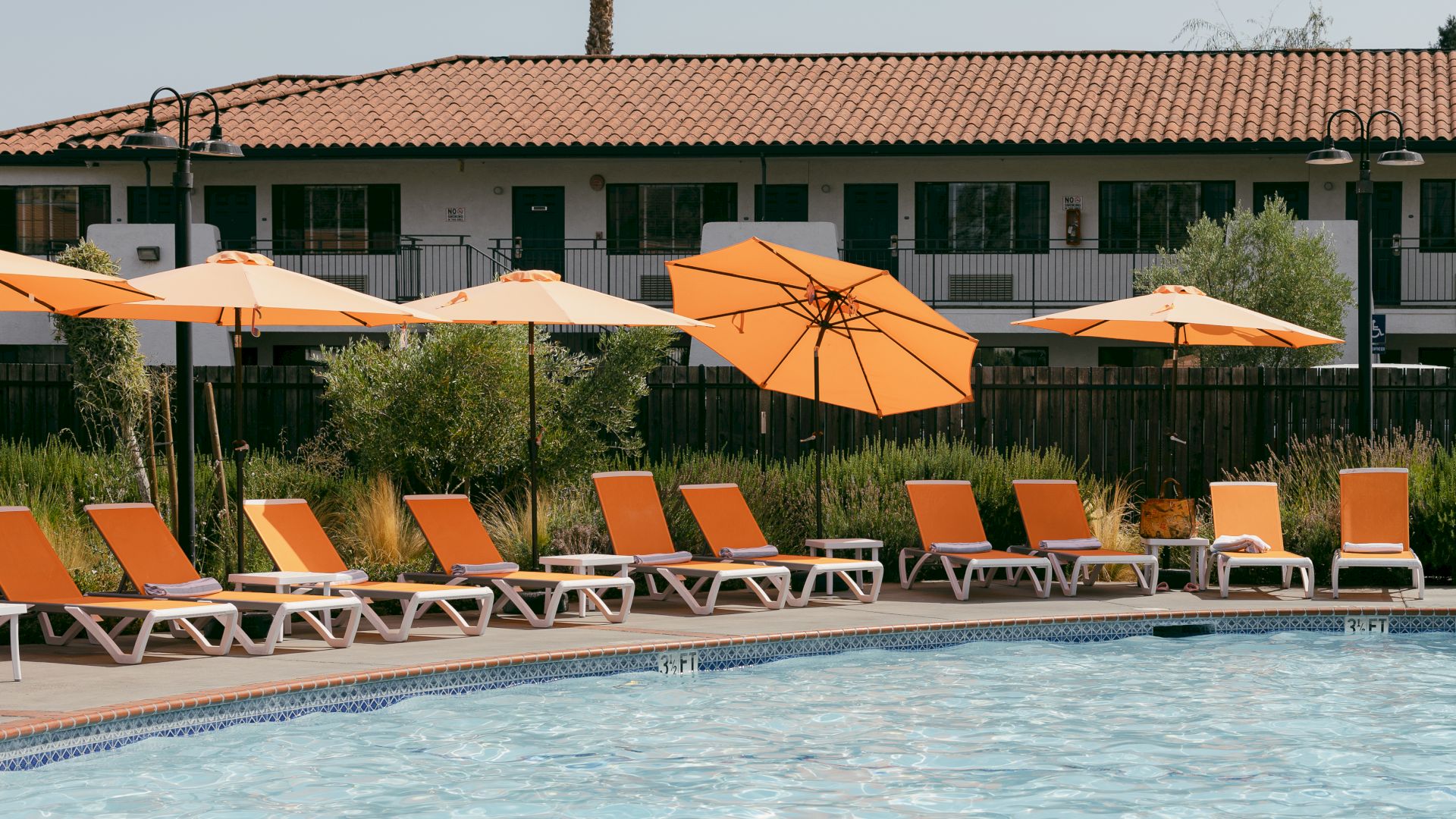 A poolside area with orange lounge chairs and umbrellas, set in front of a building with a tiled roof and green surroundings.