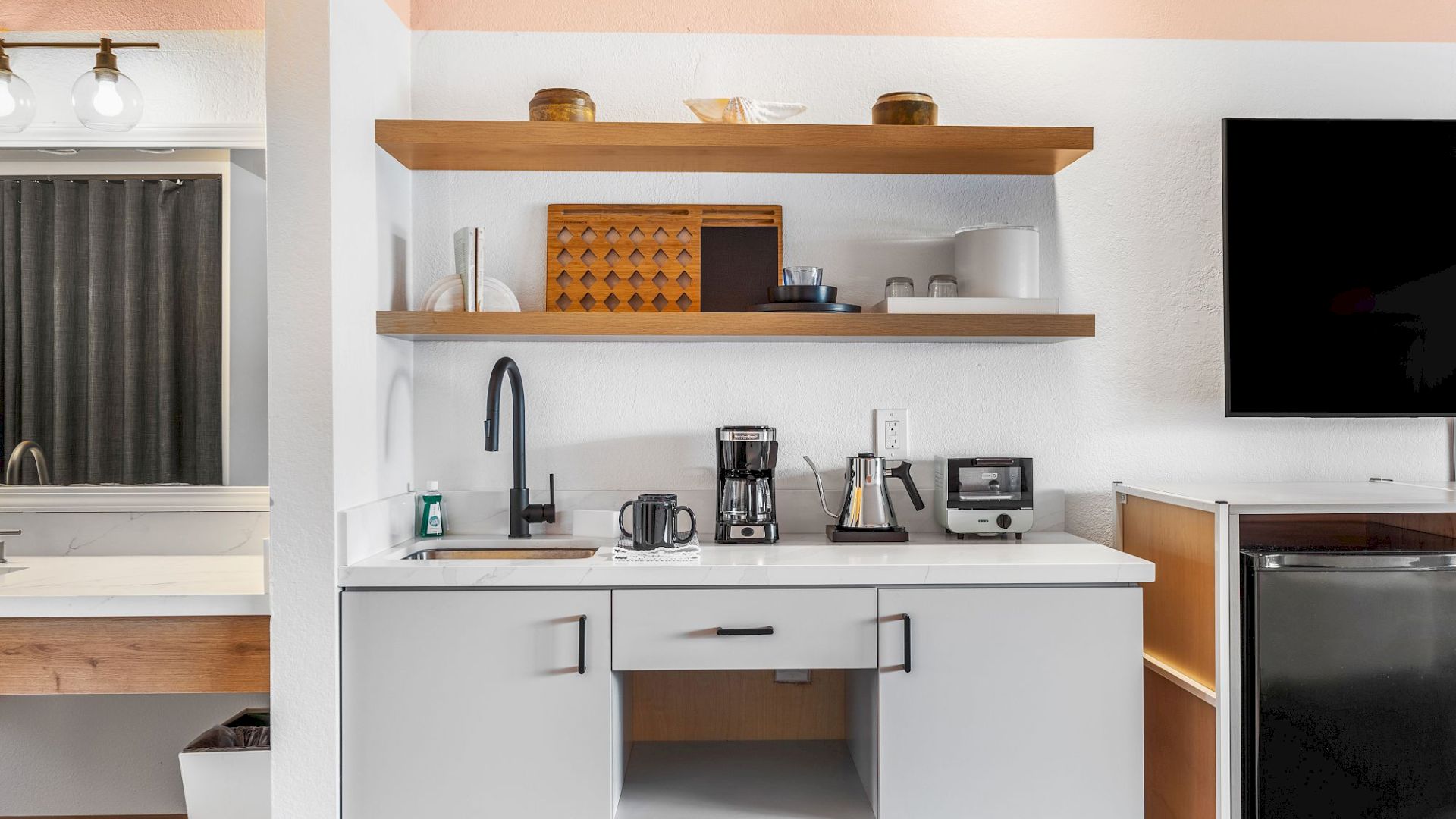 A modern kitchen counter with a coffee maker, kettle, and mugs, plus open shelves holding jars and cups above the sink.