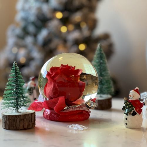 Small Christmas scene with a central crystal ball, tiny figurines (snowmen), and miniature trees on a white surface. The background features a blurred, lit Christmas tree.