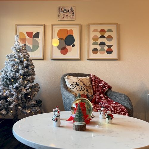A cozy living room with a frosted Christmas tree, a round coffee table, a grey armchair with a red throw, and abstract art prints on the wall, creating a festive, modern vibe.