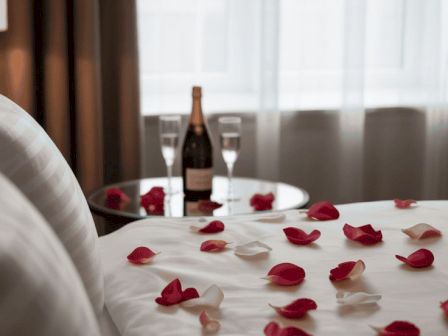 A romantic hotel bed with rose petals scattered across the sheets, a bottle of champagne and glasses by the window, aiming for a cozy, celebratory vibe.