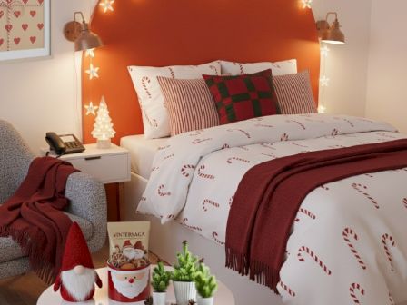 A cozy Christmas bedroom with a candy-cane theme: warm white bedding, red accents, string lights on an orange headboard, plaid pillows, and a small table with festive decor.