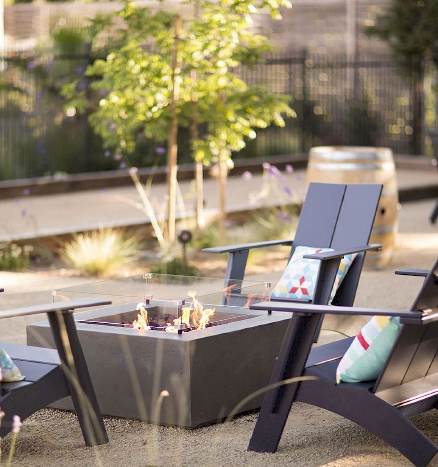 The image shows outdoor chairs around a modern fire pit with cushions and plants nearby in a sunny setting.