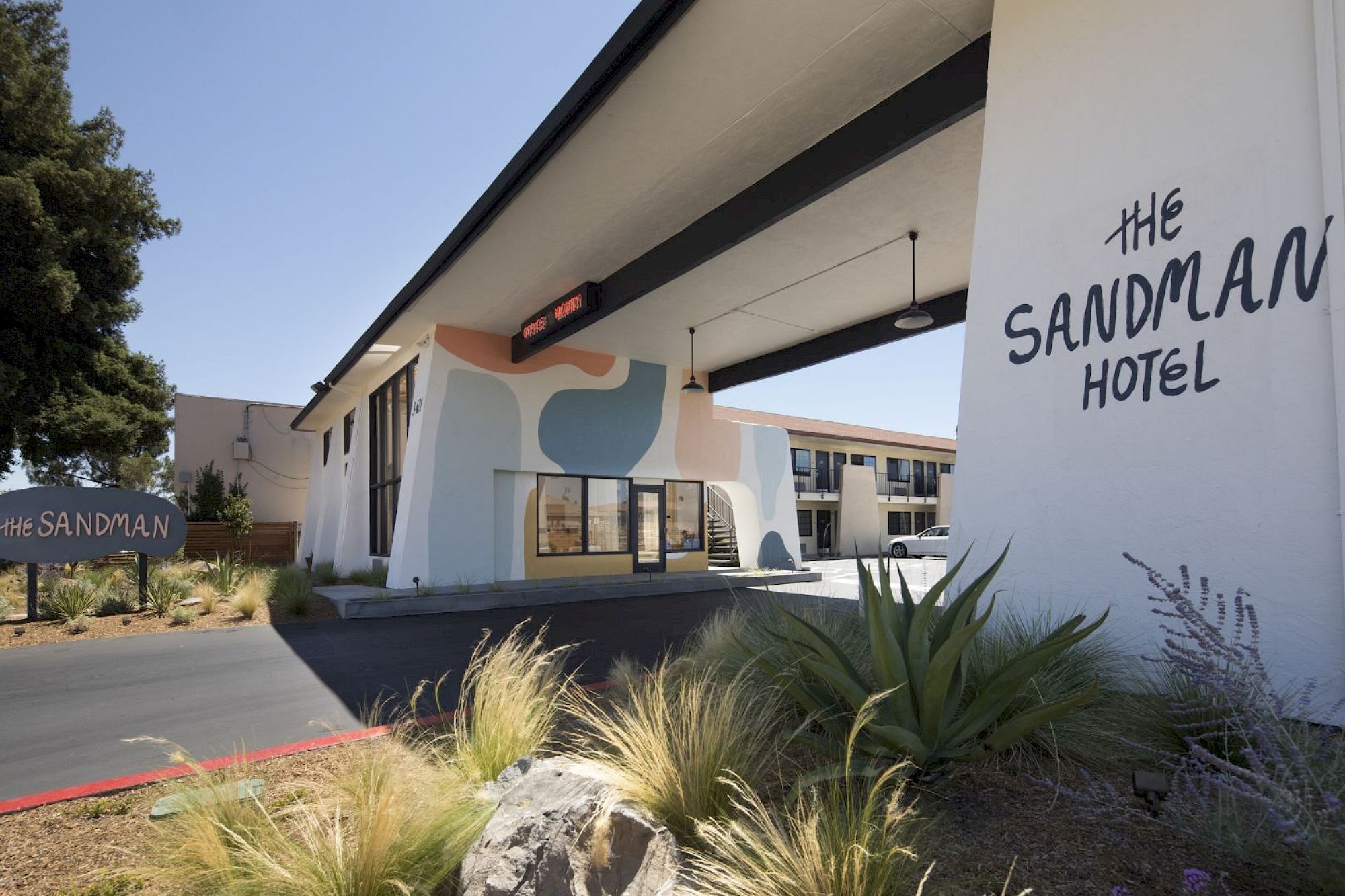 A retro motel-style building with a sign reading &ldquo;The Sandman Hotel,&rdquo; desert landscaping, and a curved entrance under a canopy. The sentence ends here.