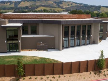 A modern single-story building with large glass windows, surrounded by a curved concrete walkway, a rust-colored steel retaining wall, and landscaped desert-style grounds.