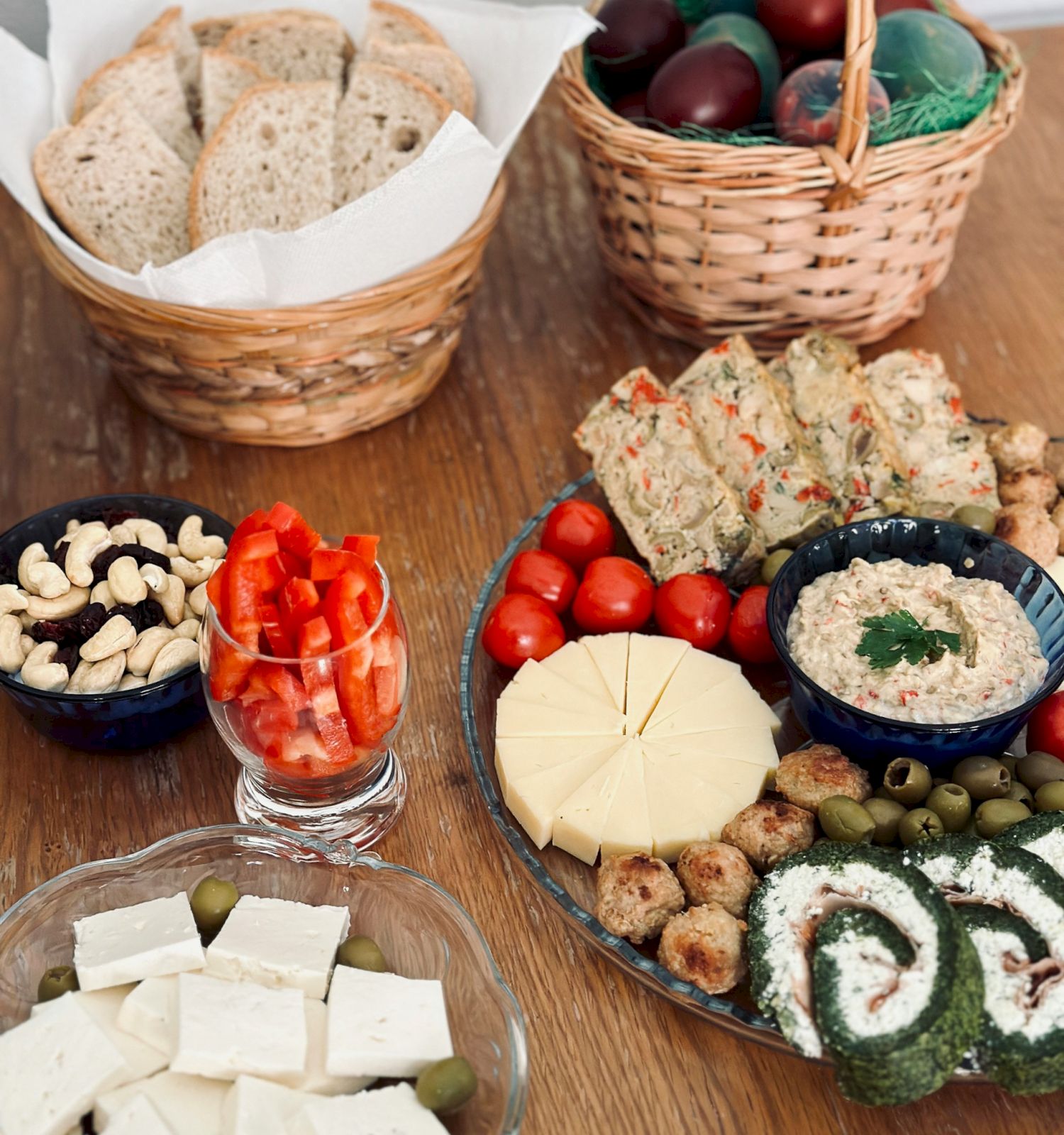 A spread of snacks: bread baskets, a basket of olives, a cheese board with assorted cheeses, crackers, nuts, grapes, cherry tomatoes, hummus, sliced veggies, and olives with greens.