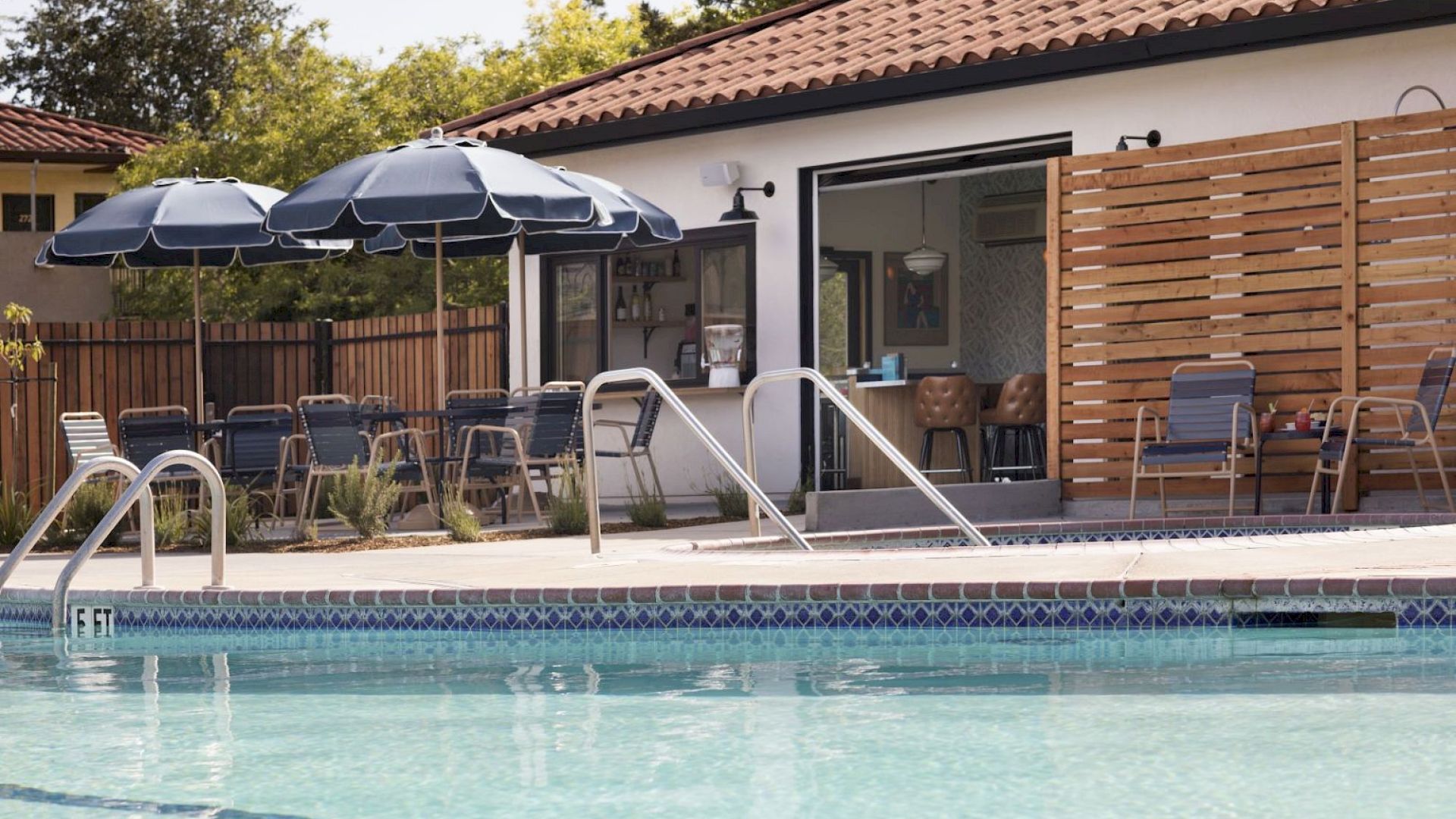 A hotel pool area with a blue-tiled swimming pool, patio seating under umbrellas, and a single-story building with a sliding door and wooden privacy screen, sunny outdoor scene.