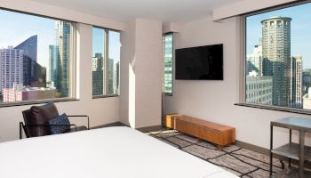 A modern hotel room with a city view, featuring large windows, a bed, an armchair, a wooden TV stand with a flat-screen TV, and a side table.