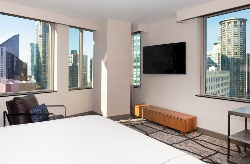 A modern hotel room with a city view, featuring large windows, a bed, an armchair, a wooden TV stand with a flat-screen TV, and a side table.