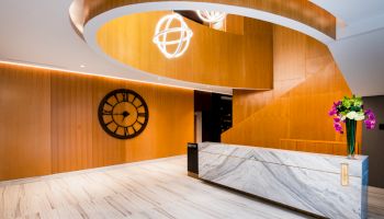 A modern reception area with a marble desk, wooden wall panels, a large wall clock, spiral staircase, and flowers on the desk, well-lit.