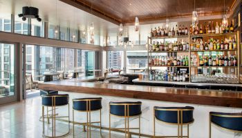 A modern bar with sleek stools, a well-stocked liquor shelf, and large windows offering a city view in the background.