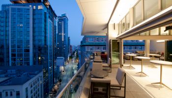 A modern rooftop terrace with tables and chairs overlooks a city street and high-rise buildings at dusk, featuring large windows and open spaces.