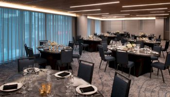 The image shows an elegantly set event or dining room with round tables, chairs, tableware, and floral centerpieces, illuminated by soft lighting.
