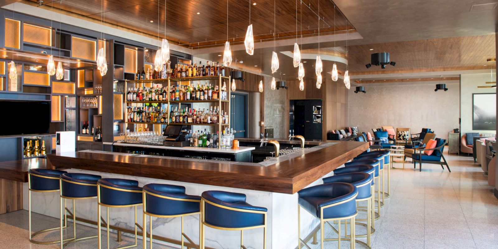 This image shows a modern bar with a wooden counter, blue bar stools, hanging lights, and a stocked bar. The seating area is visible in the background.