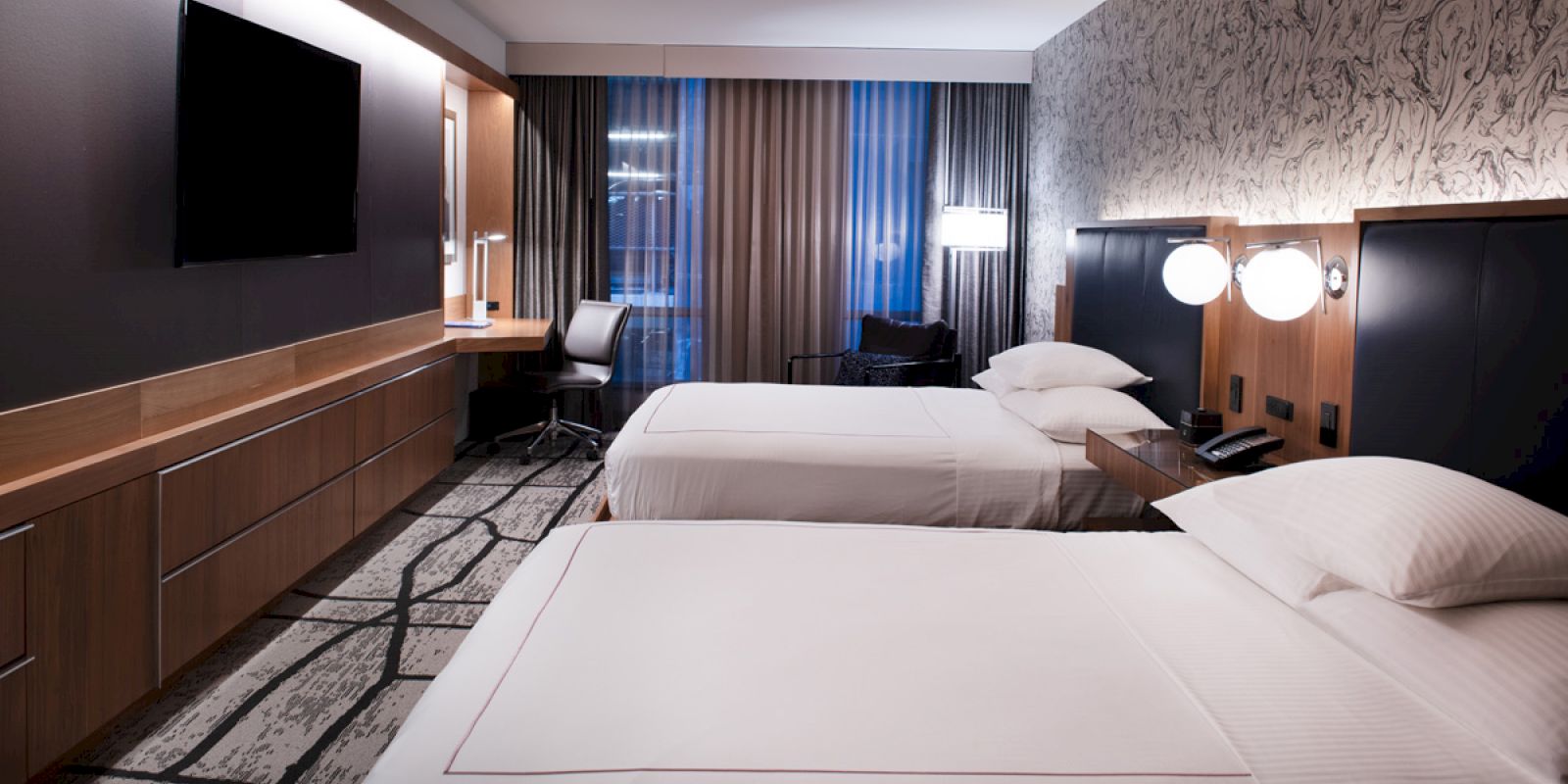 This image shows a modern hotel room with two neatly made beds, a TV, a desk and chair, lamps, and patterned carpet and wallpaper.