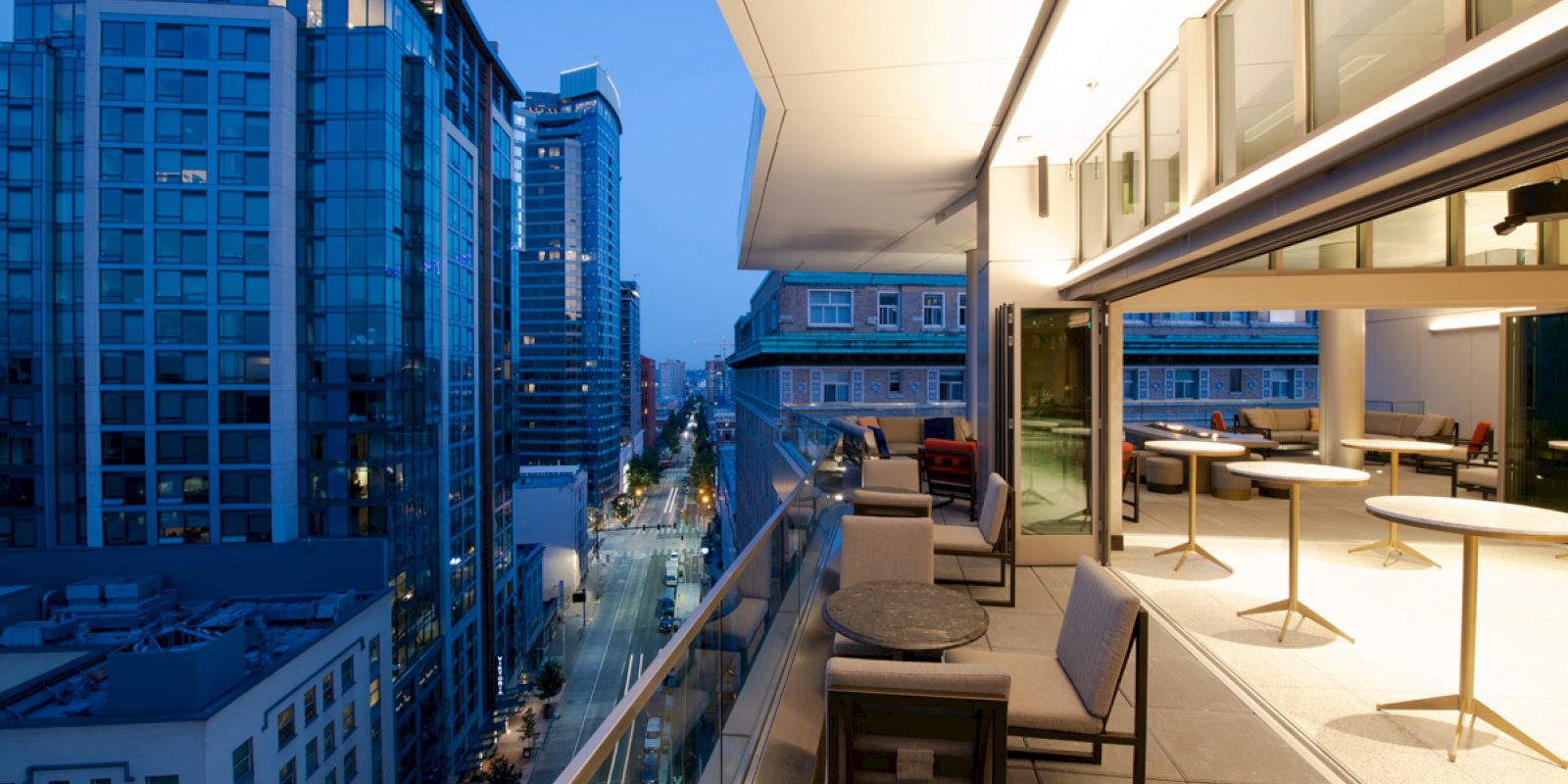 A modern balcony with seating overlooks a city street lined with tall buildings. The area is well-lit, suggesting a bustling urban environment.