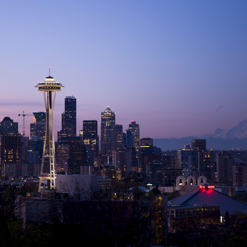This image shows the Seattle skyline at dusk, featuring the Space Needle and Mount Rainier in the background, with city lights starting to glow.