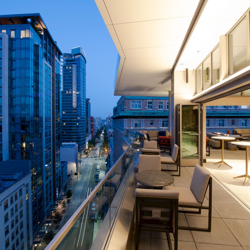 A modern balcony view: cityscape with tall buildings at dusk, featuring outdoor seating areas and tables under soft lighting, extending from indoors.
