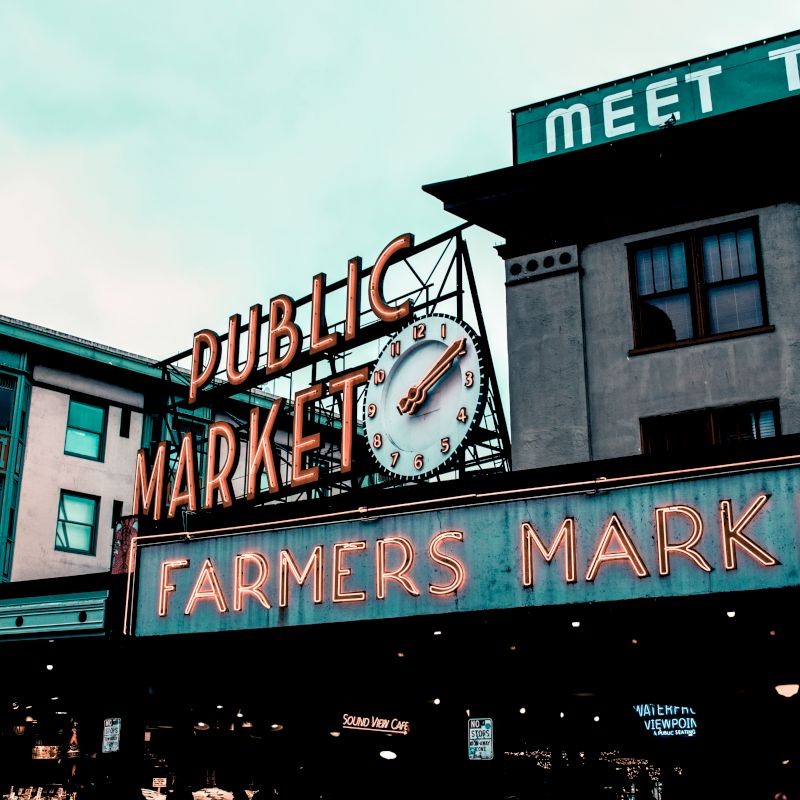 The image shows a public market building with a "Public Market" and "Farmers Market" neon sign, and a clock. The atmosphere is urban and lively.