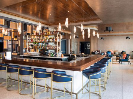 Modern bar with high chairs, hanging lights, and a well-stocked liquor display. Cozy seating area in the background.
