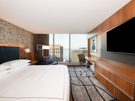 A modern hotel room with a large bed, a desk, a chair, a flat-screen TV, large windows offering a city view, and contemporary decor.