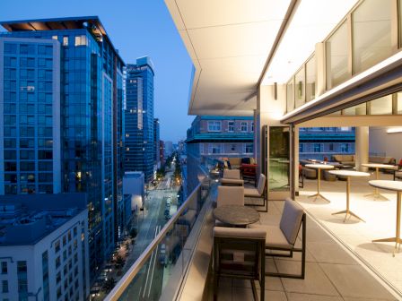 A modern cityscape view at twilight with high-rise buildings and a stylish outdoor balcony featuring seating tables and chairs, overlooking a street.