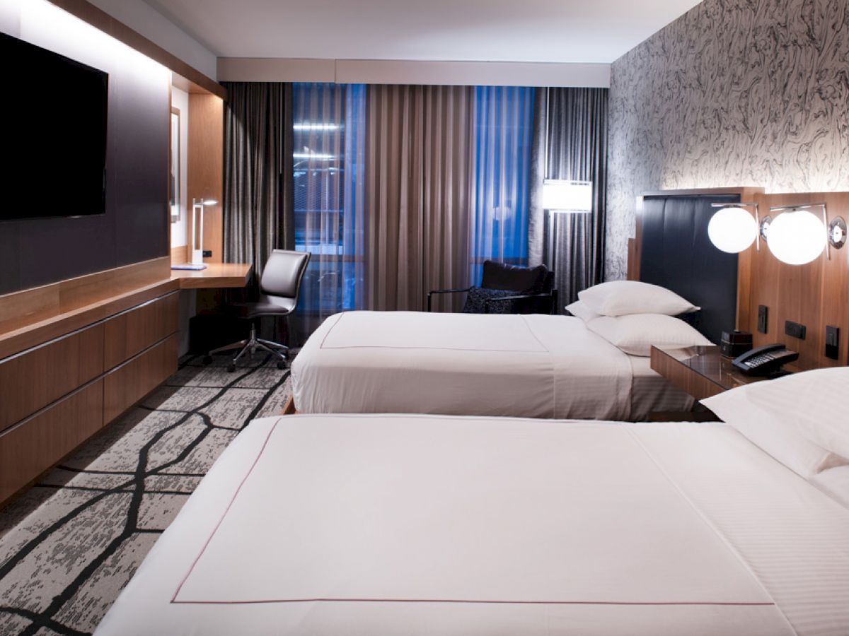 A modern hotel room with two beds, a desk and chair, a flat-screen TV, and stylish lighting and decor.