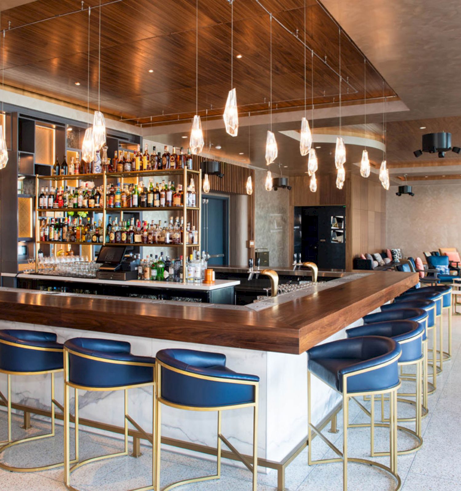 A modern bar with a stocked shelf, hanging lights, blue bar stools, and seating areas in the background, featuring contemporary decor.