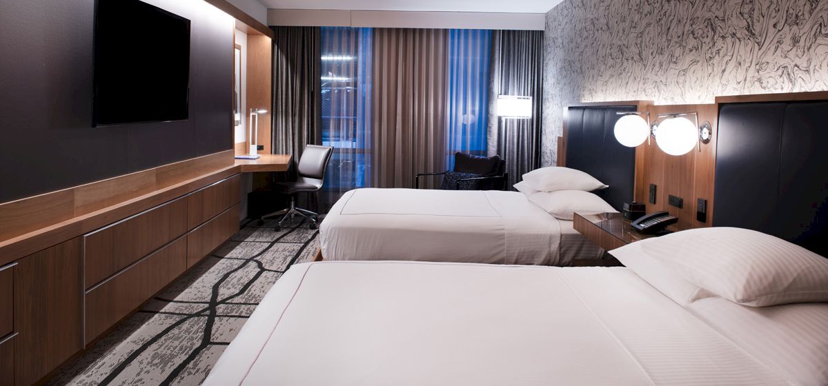 The image shows a modern hotel room with two double beds, a flat-screen TV, a work desk, and contemporary decor.
