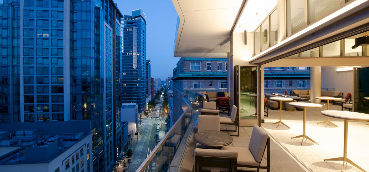 A modern cityscape view from a high-rise balcony, featuring outdoor seating, tables, and floor-to-ceiling windows, overlooking the street below.