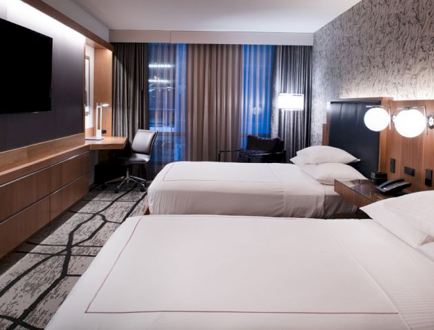 A modern hotel room with two beds, a desk with a chair, and a wall-mounted TV. There are contemporary light fixtures and patterned walls.