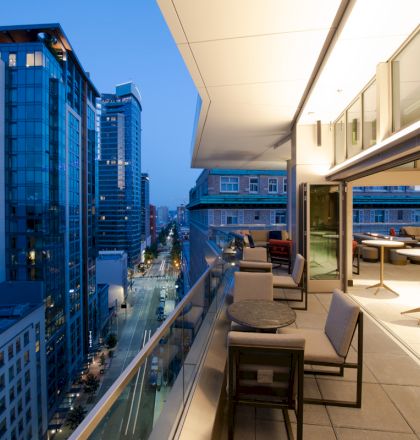 A modern urban balcony scene at dusk, featuring outdoor seating and tables, overlooking a city street lined with tall buildings and bright lights.