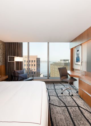This image shows a modern hotel room with a large bed, a wall-mounted TV, a desk, and a window offering a cityscape view.