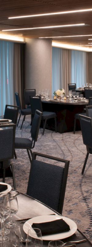 The image shows an elegantly set dining room with round tables, black chairs, and table settings, including glasses and plates, under modern ceiling lights.