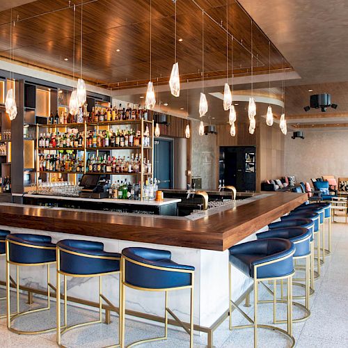 A modern bar with a wooden counter, blue stools, a wall of liquor bottles, pendant lights, and a sleek, open seating area in the background.