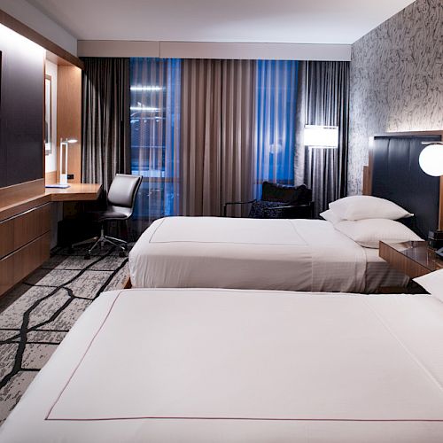 A hotel room features two double beds, a wall-mounted TV, a desk with a chair, modern lighting, and a patterned wall and carpet.