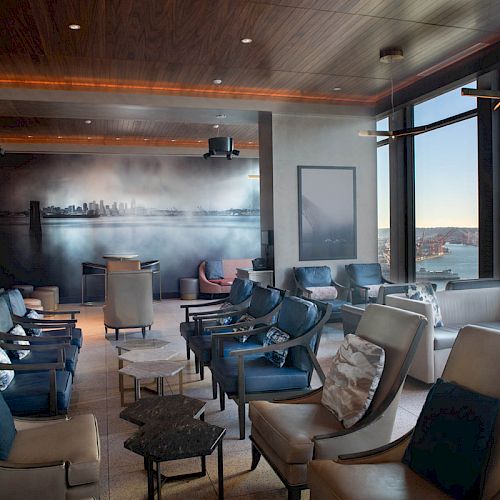 A modern lounge with plush seating, large windows offering a scenic view of the water and a ferris wheel, and a city skyline mural on the wall.