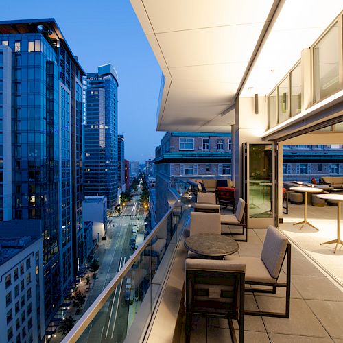 A modern cityscape at dusk with tall buildings and a sleek balcony featuring outdoor seating and tables overlooking the illuminated street.