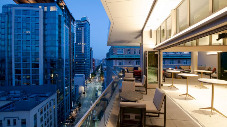 A modern high-rise balcony at dusk with seating area, tables, and city street view below, lined with other tall buildings.