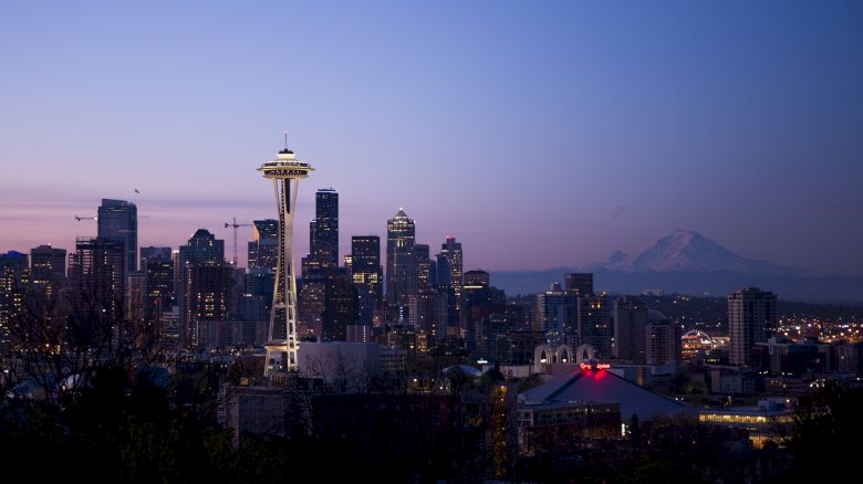 The image depicts a city skyline at dusk with the iconic Space Needle prominently visible and Mount Rainier in the background.