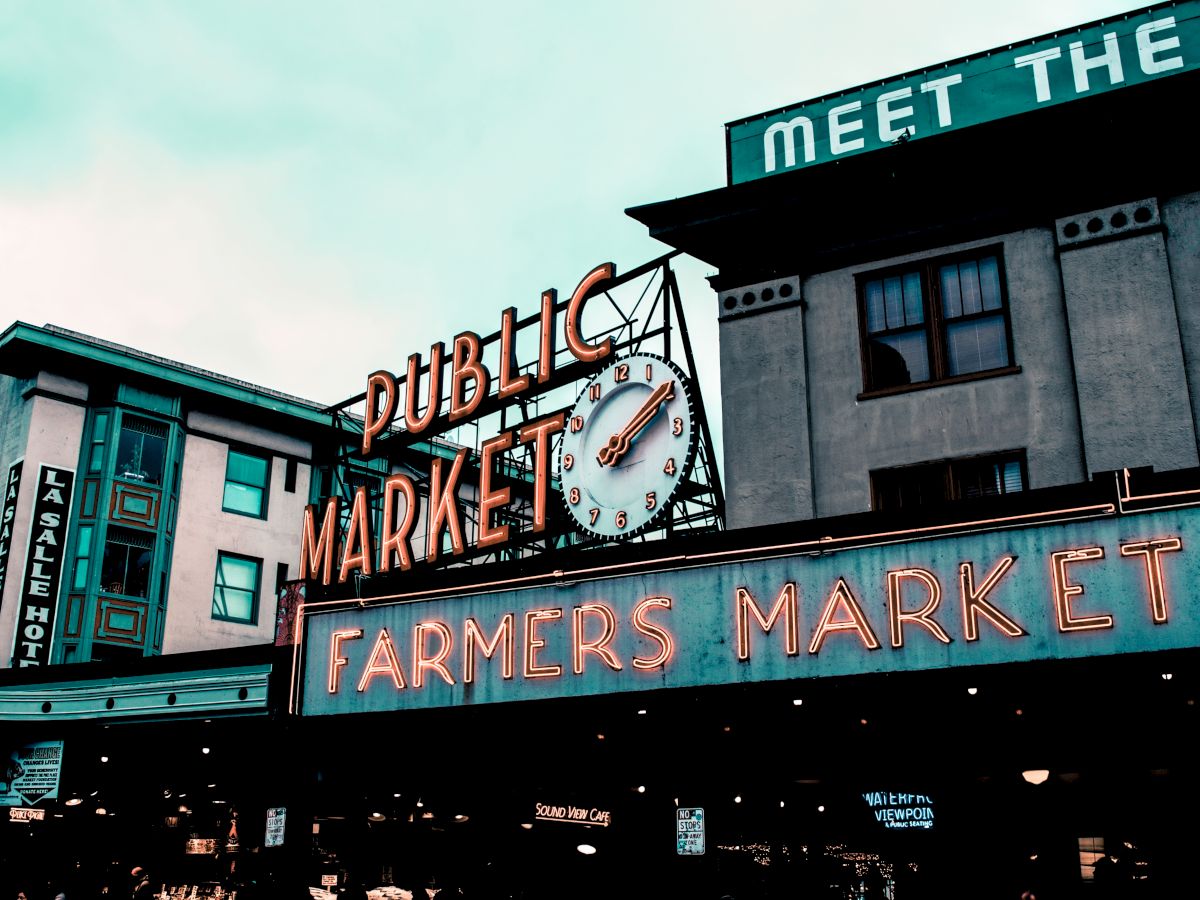 The image shows an iconic public market with neon signs reading "Public Market" and "Farmers Market" along with adjacent buildings and a clock.
