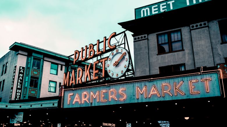 The image shows an iconic public market with neon signs reading "Public Market" and "Farmers Market" along with adjacent buildings and a clock.
