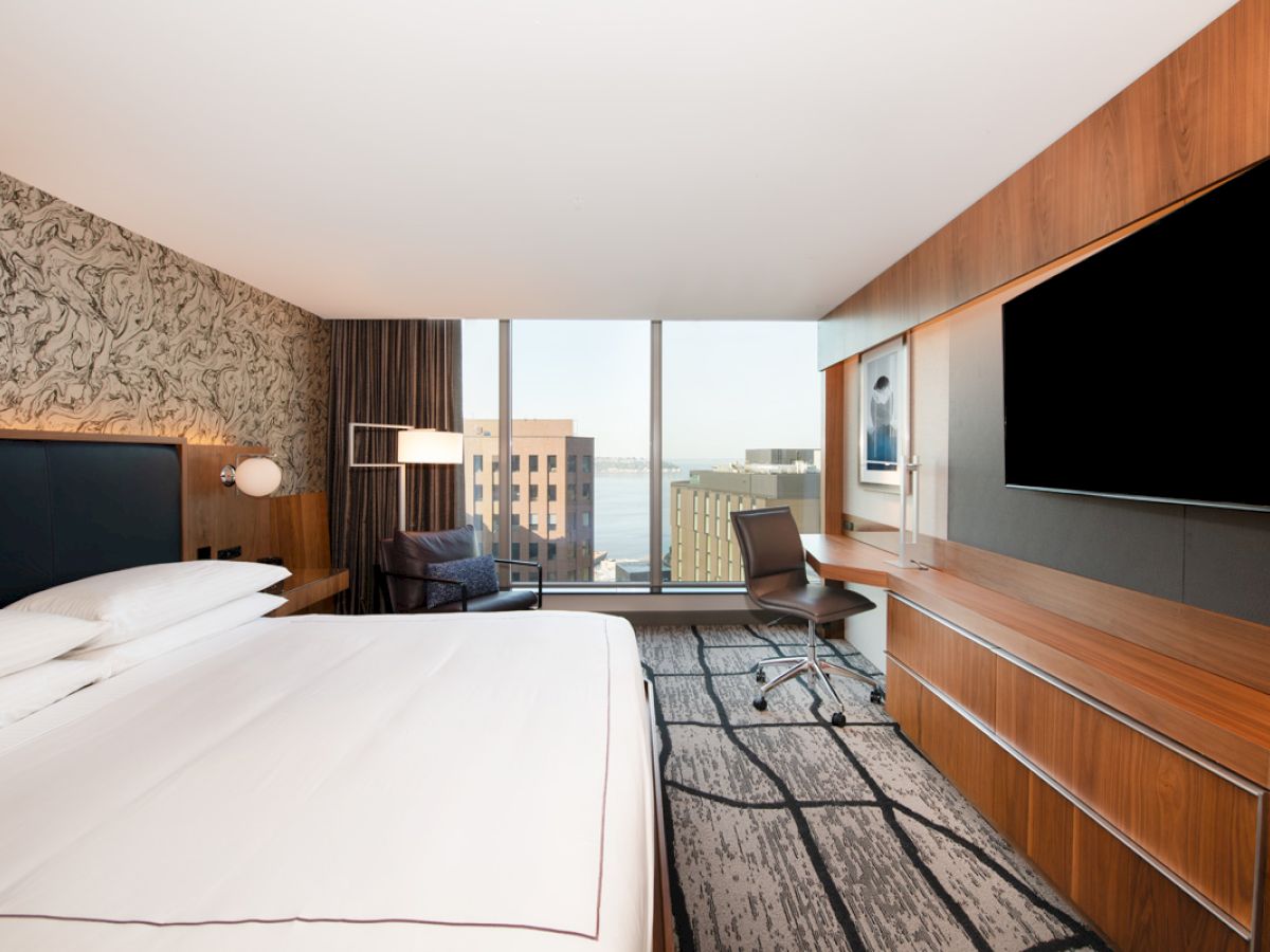 A modern hotel room with a king-size bed, desk, chair, large TV, and a window view of city buildings and water.