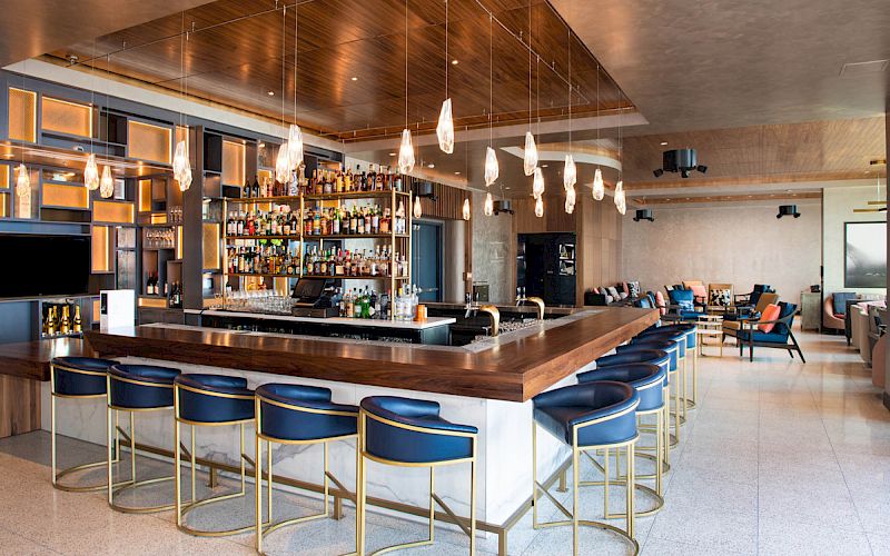 This image shows a modern bar with blue bar stools, pendant lighting, a stocked liquor display, and a lounge area in the background.